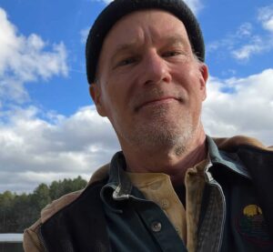 man in outdoors attire and beenie cap with blue sky background and clouds, lake and trees
